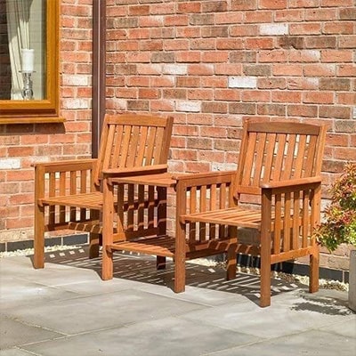 Two wooden connected garden chairs/benches with table in the middle., outside a brick house.